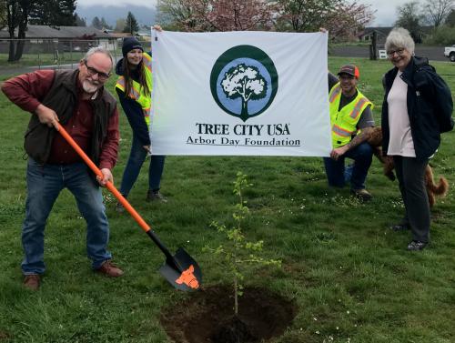 Newly planted Ginkgo tree with man holding a shovel with the Tillamook City logo. People in the background hold a Tree City USA / Arbor Day Foundation banner.